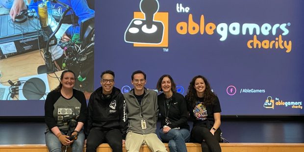 AbleGamers Peer Counseling staff members Jessie and Andy along with three Shepherd Center staff members sitting the edge of a stage, a large screen in back of them shows a slide with the AbleGamers logo from a presentation about the AbleGamers and Shepherd Center Partnership