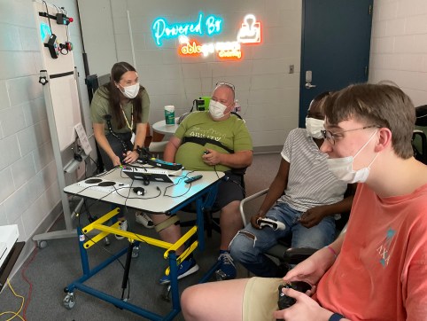 three men playing video games together using several adaptive gaming devices a young woman who is an occupational therapist is watching photo provided by Ability KC a Powered by AbleGamers partner