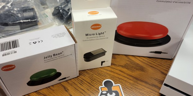 AbleGamers partnerships - Craig Hospital 3 boxes, 3 different switches green jelly bean switch, microLight and a Big Red sitting on a wooden table top with an ablegamers sticker in the center