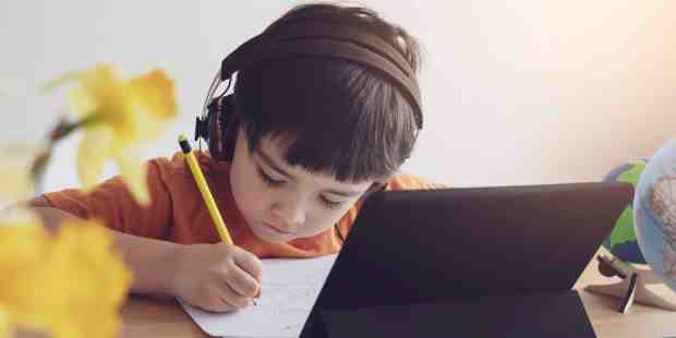 young man with a headset on, writing on paper with an iPad propped up in front of him.