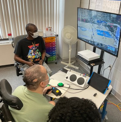 Two people watching someone play a game. There is an xbox adaptive controller with various switches plugged in. The player is holding a joycon.