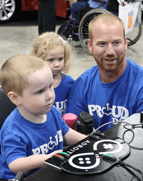 AbleGamers-Splash1 an image of three people, one man kneeling next to a young boy who is sitting in a chair and a young girl standing in the background