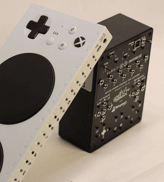 An AbleGamers Adroit Switchblade, a black box with silver switch ports all over it. Leaning on the Adroit is a Microsoft XAC, a white candybar shapped device with ports on the back and a large black button.