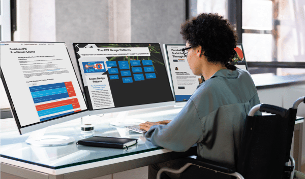 a young woman sitting at a desk facing three computer screens. On the computer screens are the AbleGamers apx course information. Support the DPAD Initiative, host a fundraiser