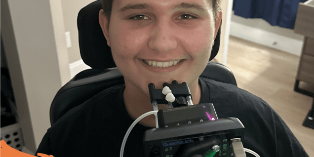 A photo of Carson, he has short dark hair, brown eyes and wearing a black shirt. He is smiling and in front of him is a QuadStick adaptive controller. He can play video games again.