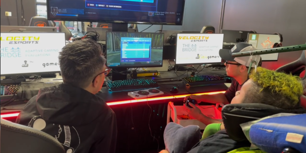 Andy and 2 gamers, one in a powered wheelchair, playing rocket league. More game stations are in the background.