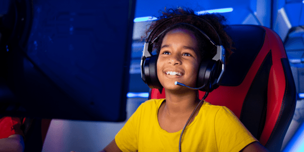 A child wearing a gaming headset sitting in a gaming chair in front of a computer screen playing summer video games..