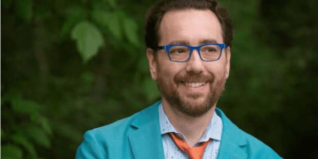 a smiling Mark Barlet, founder and Executive Director of the AbleGamers wearing a teal suit jacket, light blue shirt, orange tie and royal blue glasses