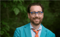 a smiling Mark Barlet, founder and Executive Director of the AbleGamers wearing a teal suit jacket, light blue shirt, orange tie and royal blue glasses
