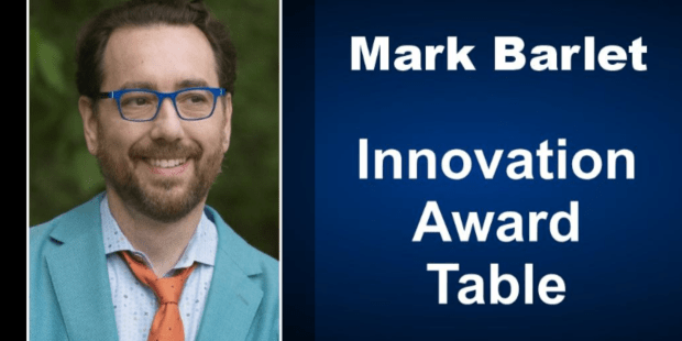Mark Barlet head shot, he is wearing royal blue glasses, a teal suit jacket, blue dress shirt and AbleGamers orange tie. Next to Mark's photo is written Mark Barlet Innovation Award Table