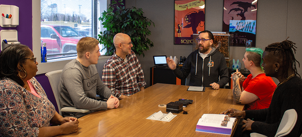 Mark Barlet, middle aged white male, brown hair and beard. Sitting at the head of a table with other AbleGamers team members. They are listening to Mark