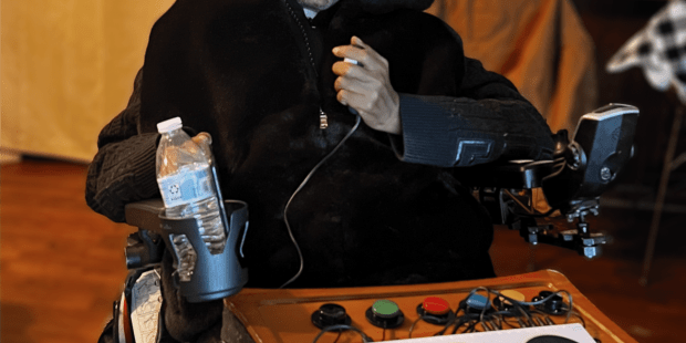 Roland, an African=America male with a black hoodie. He is in a wheelchair holding a joystick in his hand. In front of him is an XAC and switches