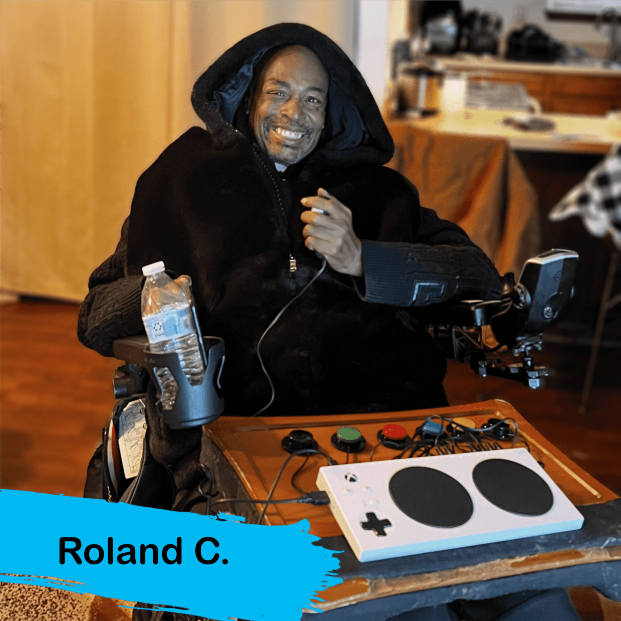 Roland, an African=America male with a black hoodie. He is in a wheelchair holding a joystick in his hand. In front of him is an XAC and switches