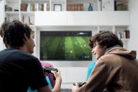 two teen boys playing a video game, one is wearing a brown hooded sweatshirt the other a black t-shirt. They are talking and smiling. The importance of connecting with others.
