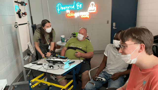 three men playing video games together using several adaptive gaming devices a young woman who is an occupational therapist is watching photo provided by Ability KC a Powered by AbleGamers partner